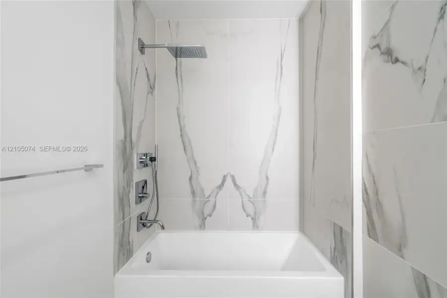 Bathtub featuring large-format veined tile surround, overhead rain shower head, handheld shower wand, and polished chrome fixtures