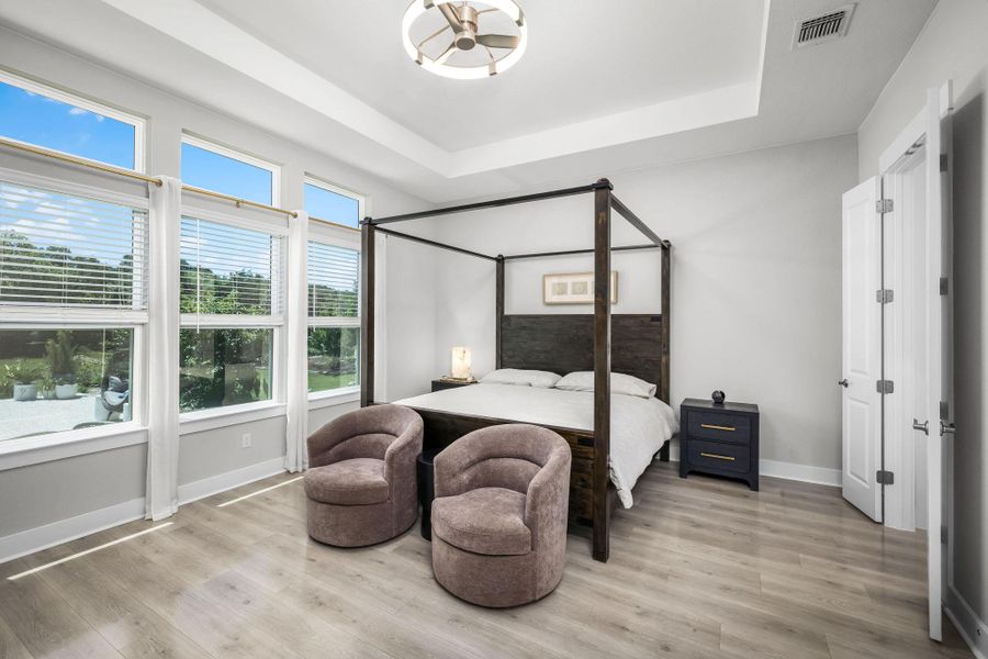 Sun-filled bedroom featuring a tray ceiling, elegant wood flooring, and extensive windows that frame the lush outdoor scenery.