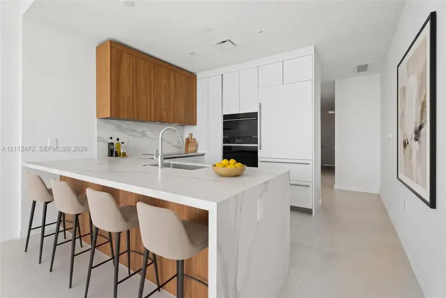 Kitchen featuring waterfall-edge stone island with integrated sink, flat-panel wood cabinetry, white floor-to-ceiling cabinetry, stone backsplash, and built-in wall oven. VIRTUALLY STAGED