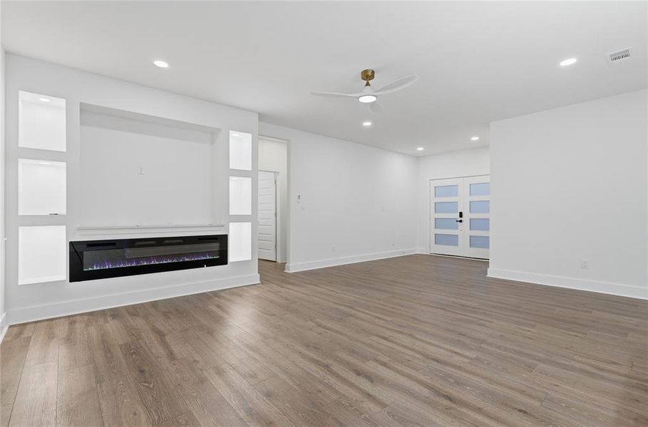 Spacious open-concept room featuring wood-finish flooring, a built-in media center with shelving and an electric fireplace, a modern ceiling fan, recessed lighting, and a double entry door with frosted glass panels