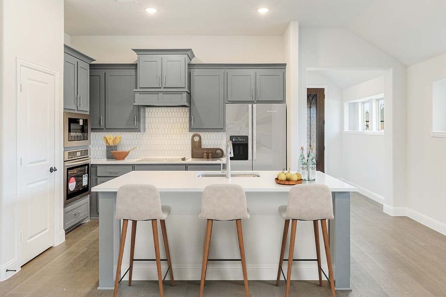 Kitchen with gray cabinets, white island with three bar stools, tile backsplash, and stainless steel appliances