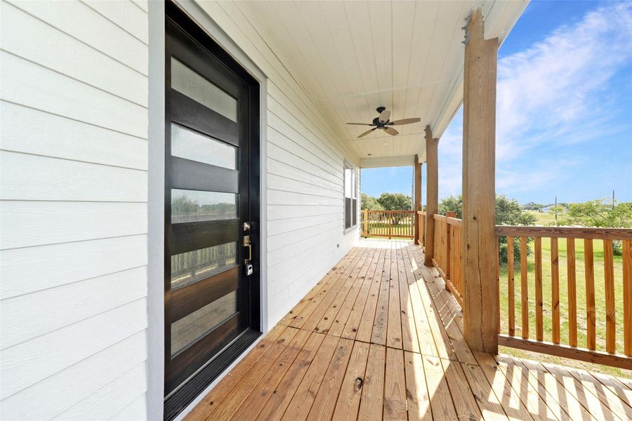 This photo features a spacious wooden deck with a modern black door and glass panels. The deck has a ceiling fan, wooden railings, and overlooks a green landscape, offering a serene outdoor space.