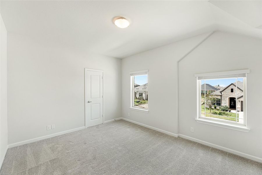 Empty bedroom with vaulted ceiling, gray carpet, and two windows overlooking neighboring homes