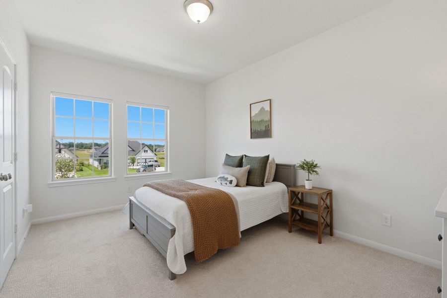 Bright and cozy bedroom with a large window offering plenty of natural light and a view of the neighborhood. Features include a neatly made bed, neutral carpeting, and simple decor.