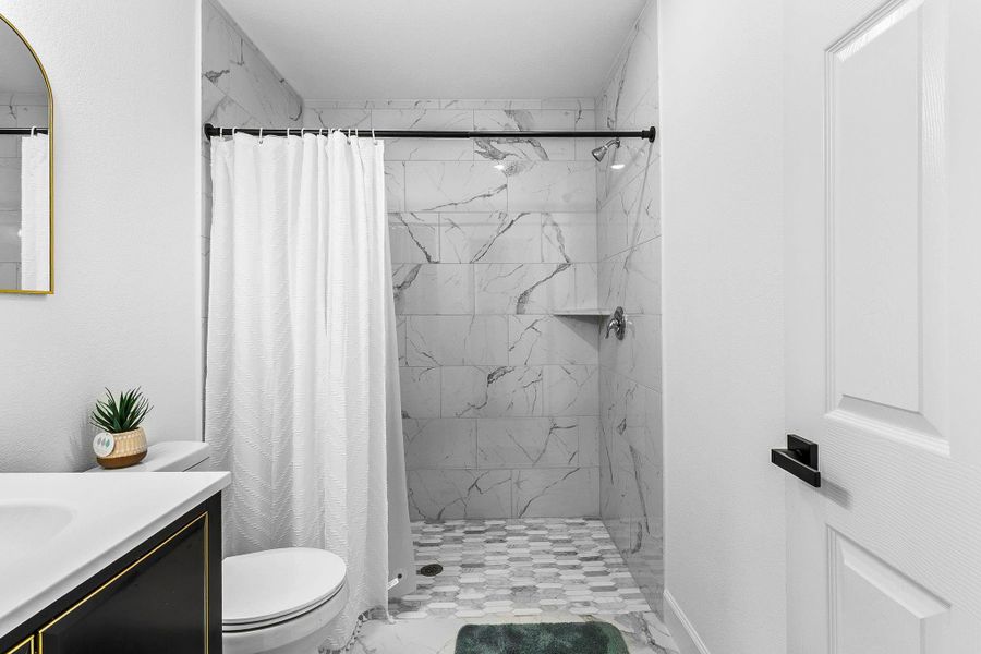 This bathroom features a modern design with marble-style tile walls and a walk-in shower. It includes a sleek vanity with a decorative mirror and a small potted plant for a touch of greenery. The space is bright and clean, highlighting a minimalist aesthetic.