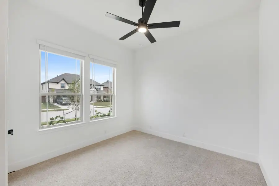 Empty bedroom with carpet flooring, black ceiling fan, and two windows overlooking a neighborhood street