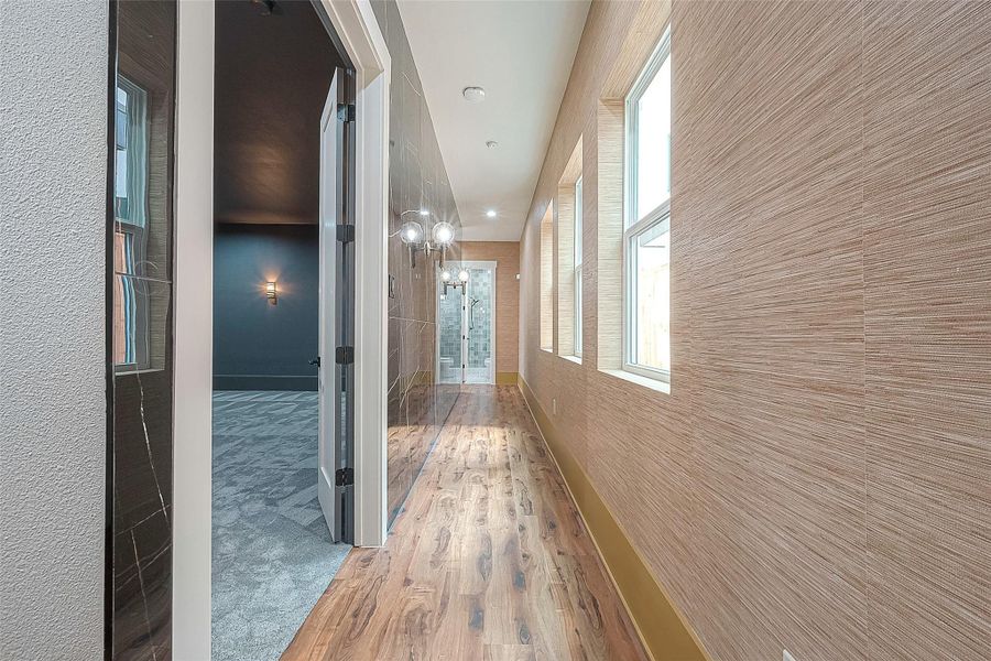 This is a modern hallway featuring natural wood flooring, textured walls, and ample natural light from the row of windows on one side. A clean-lined, glass door leads to another room, enhancing the contemporary feel of the space. This is a modern hallway featuring natural wood flooring, textured walls, and ample natural light from the row of windows on one side. A clean-lined, glass door leads to another room, enhancing the contemporary feel of the space.