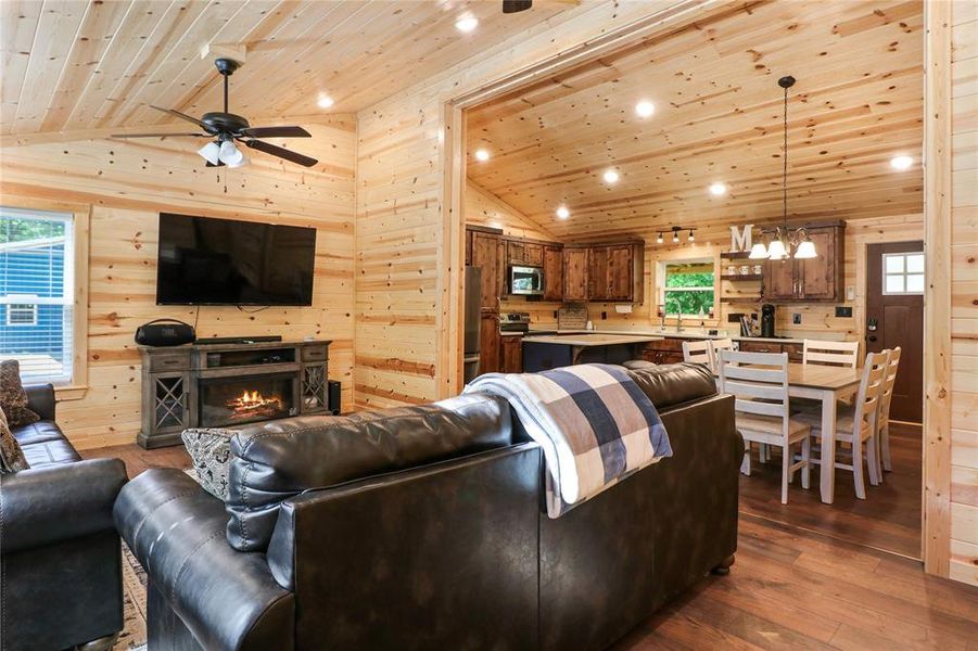 Living room with ceiling fan with notable chandelier, wooden walls, wood-type flooring, high vaulted ceiling, and wood ceiling
