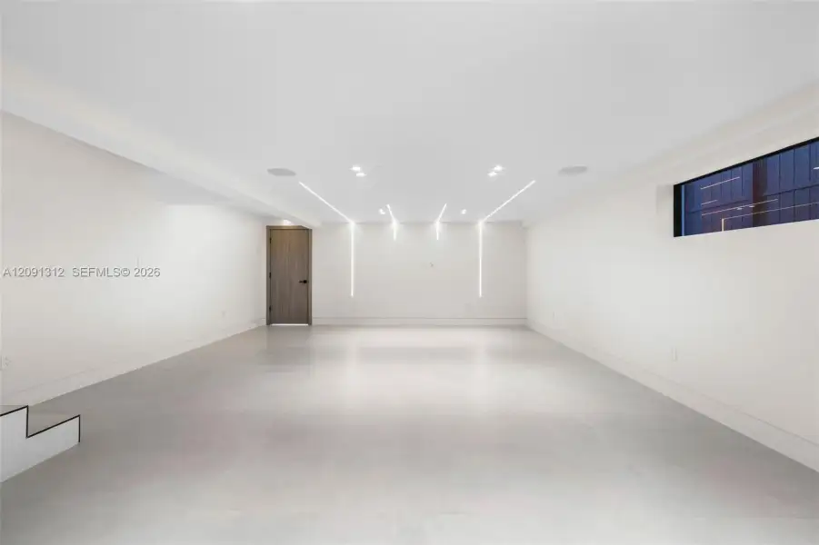 Open interior featuring expansive smooth-surface flooring, integrated recessed linear LED accent lighting, minimalist high-ceiling design, and a solid-core wood-finish door with matte black hardware Open interior featuring expansive smooth-surface flooring, integrated recessed linear LED accent lighting, minimalist high-ceiling design, and a solid-core wood-finish door with matte black hardware