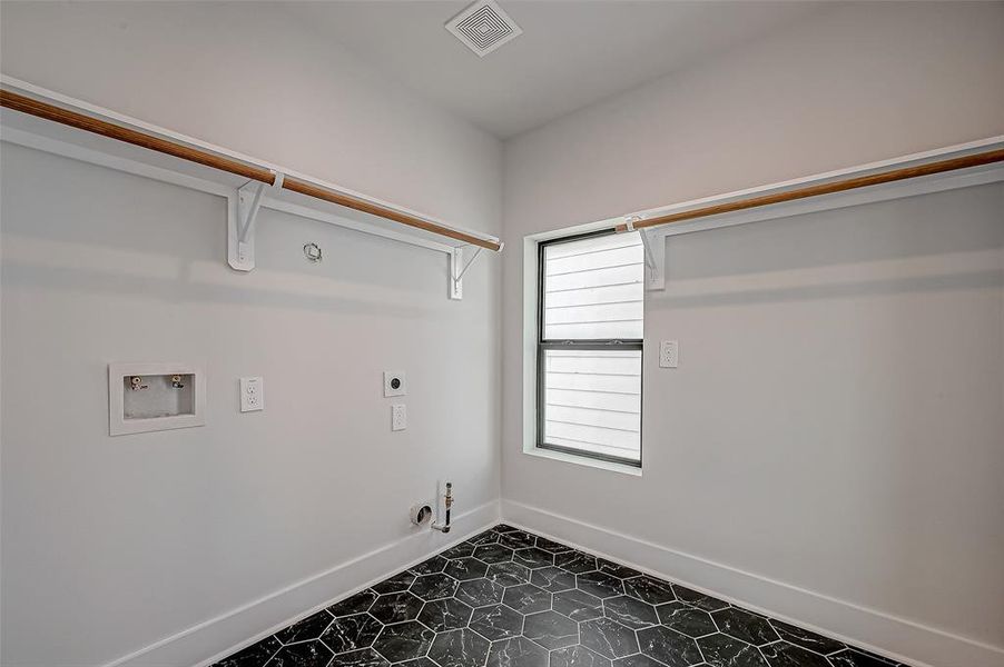 This photo shows a bright laundry room with hookups for a washer and dryer, featuring white walls, a window for natural light, and black hexagonal tile flooring. There are two wooden shelves for storage.