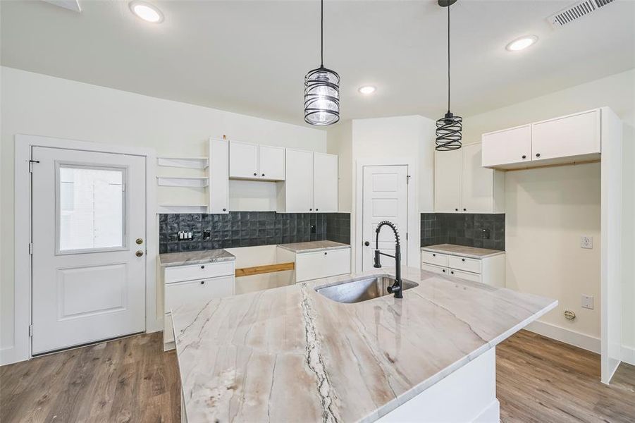 Kitchen featuring a large island with a composite countertop and undermount sink, white cabinetry with black hardware, dark square tile backsplash, and wood-finish flooring Kitchen featuring a large island with a composite countertop and undermount sink, white cabinetry with black hardware, dark square tile backsplash, and wood-finish flooring