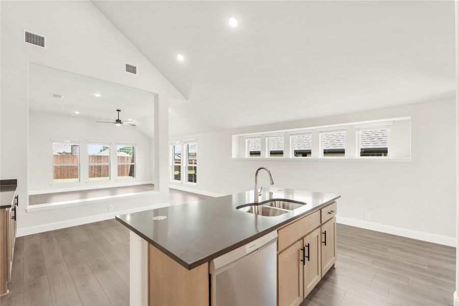 Kitchen island with dark countertop, stainless steel sink, and light wood cabinets, open to living area with vaulted ceiling. Kitchen island with dark countertop, stainless steel sink, and light wood cabinets, open to living area with vaulted ceiling.
