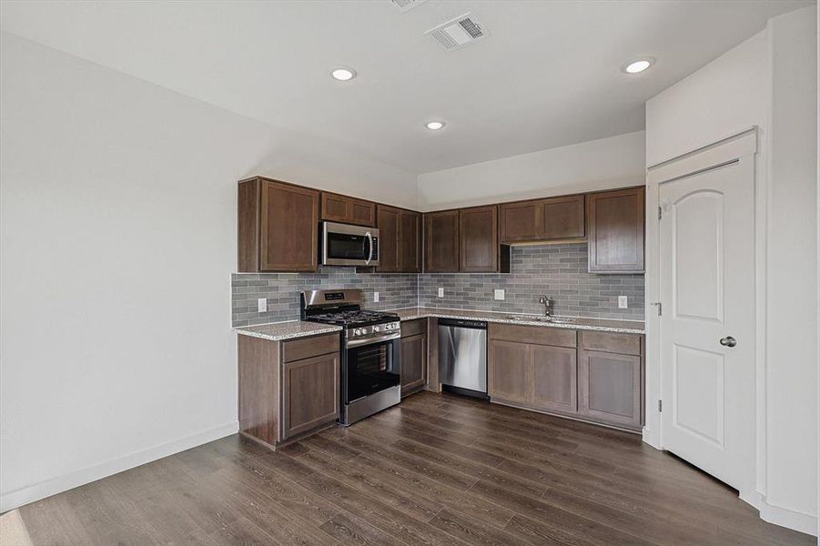 Kitchen with stainless steel appliances, dark brown cabinetry, backsplash, light stone counters, and dark wood-type flooring