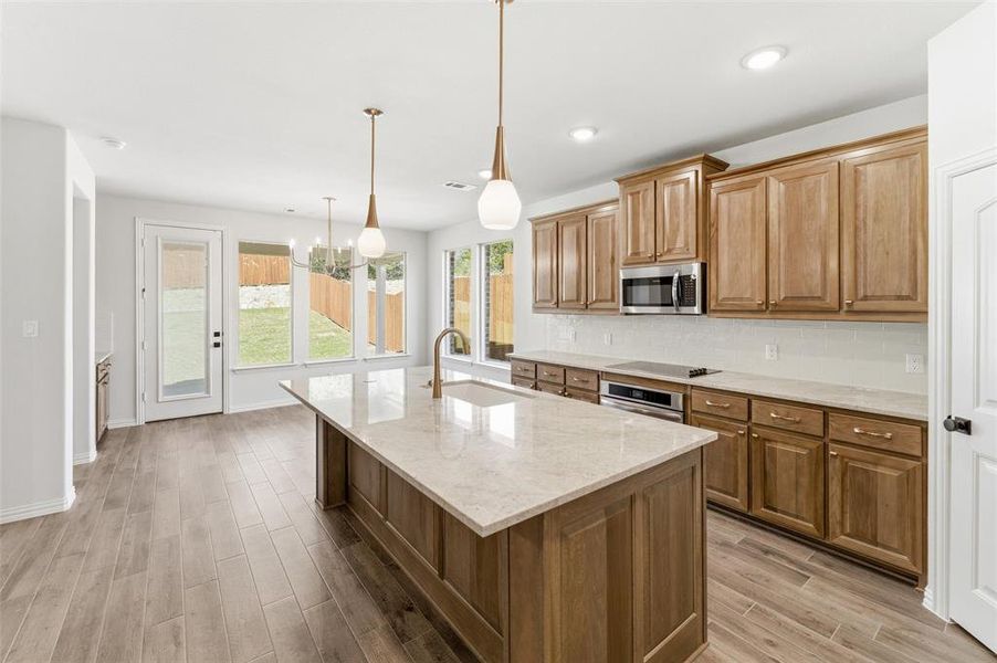 Kitchen featuring tasteful backsplash, wood finish floors, light stone counters, hanging lights, and a center island with sink