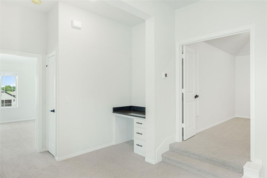 Carpeted hallway with built-in desk nook, white walls, and doors leading to adjacent rooms