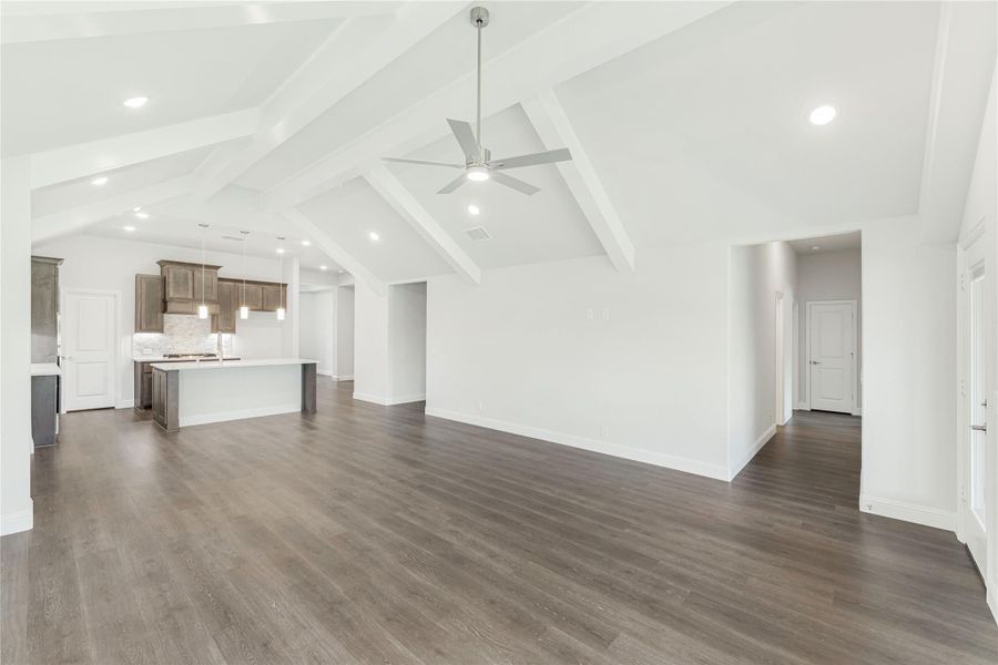 Open living area with vaulted beamed ceiling, ceiling fan, dark hardwood floors, and kitchen with island in background. Open living area with vaulted beamed ceiling, ceiling fan, dark hardwood floors, and kitchen with island in background.