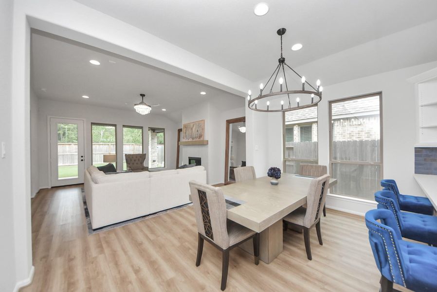 This spacious open-concept living and dining area features light wood flooring, large windows for natural light, and modern lighting fixtures. It includes a cozy living room with a view of the backyard and an elegant dining space adjacent to a stylish kitchen with blue bar stools.