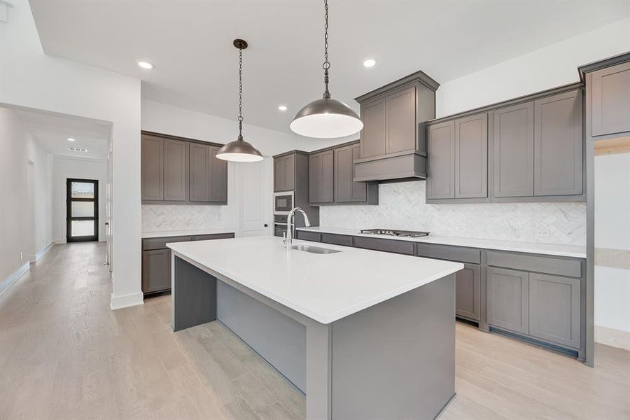 Kitchen with decorative backsplash, an island with sink, light wood-style floors, hanging light fixtures, and light stone countertops Kitchen with decorative backsplash, an island with sink, light wood-style floors, hanging light fixtures, and light stone countertops