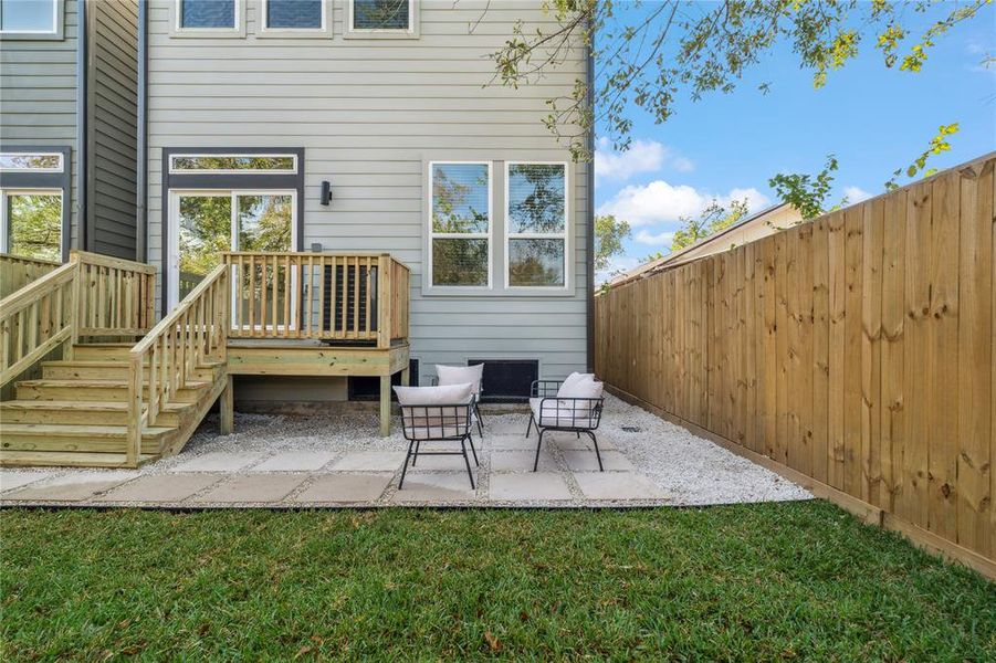 Charming backyard space with a cozy deck, seating area on a paved patio, and a fenced yard for privacy, perfect for outdoor relaxation or entertaining.