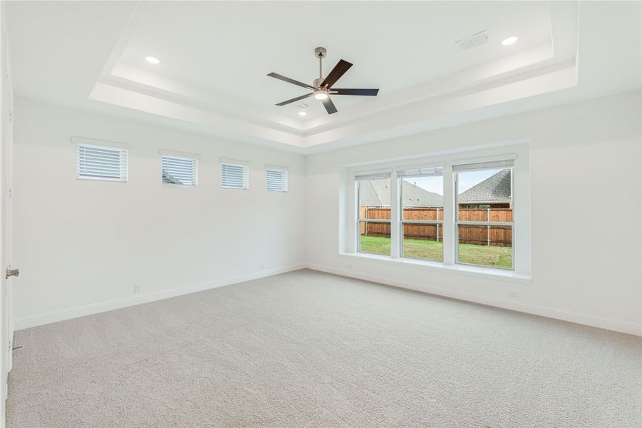 Spacious empty bedroom with tray ceiling, ceiling fan, carpet flooring, and large windows overlooking a backyard fence.