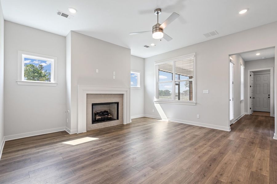 Wood-finish flooring throughout, featuring a fireplace with a white mantel, multiple windows, and a ceiling fan with integrated lighting