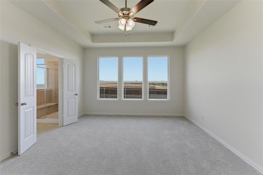 Spacious carpeted room featuring a ceiling fan with light fixture, tray ceiling, and three large windows Spacious carpeted room featuring a ceiling fan with light fixture, tray ceiling, and three large windows