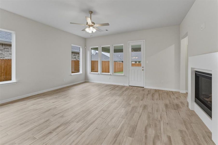 Spacious room featuring light wood-finish flooring, multiple windows, a ceiling fan with integrated lighting, a glass-paned exterior door, and a built-in fireplace with a white surround