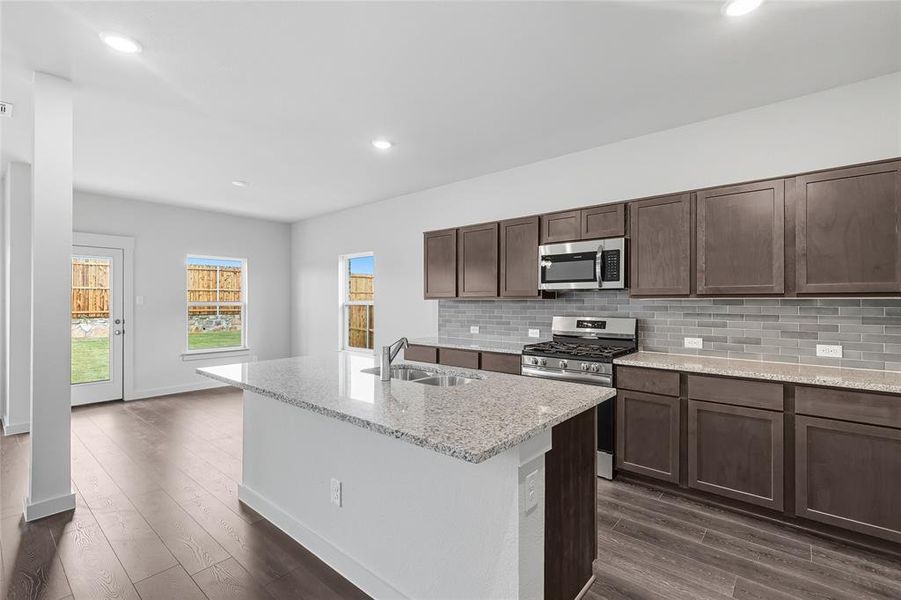 Kitchen with tasteful backsplash, light stone countertops, dark brown cabinetry, an island with sink, and recessed lighting Kitchen with tasteful backsplash, light stone countertops, dark brown cabinetry, an island with sink, and recessed lighting