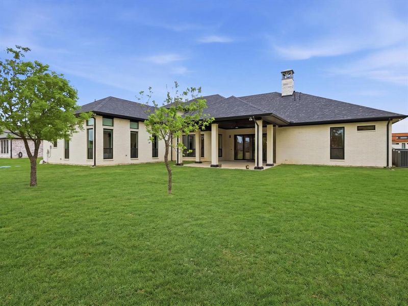 Expansive rear exterior featuring a covered patio with dark columns, a stone chimney, a dark shingle roof, and light-colored brick siding
