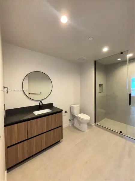 Bathroom featuring a wood-finish floating vanity with a dark countertop and integrated sink, circular wall-mounted mirror, matte black faucet, and a walk-in glass shower enclosure Bathroom featuring a wood-finish floating vanity with a dark countertop and integrated sink, circular wall-mounted mirror, matte black faucet, and a walk-in glass shower enclosure