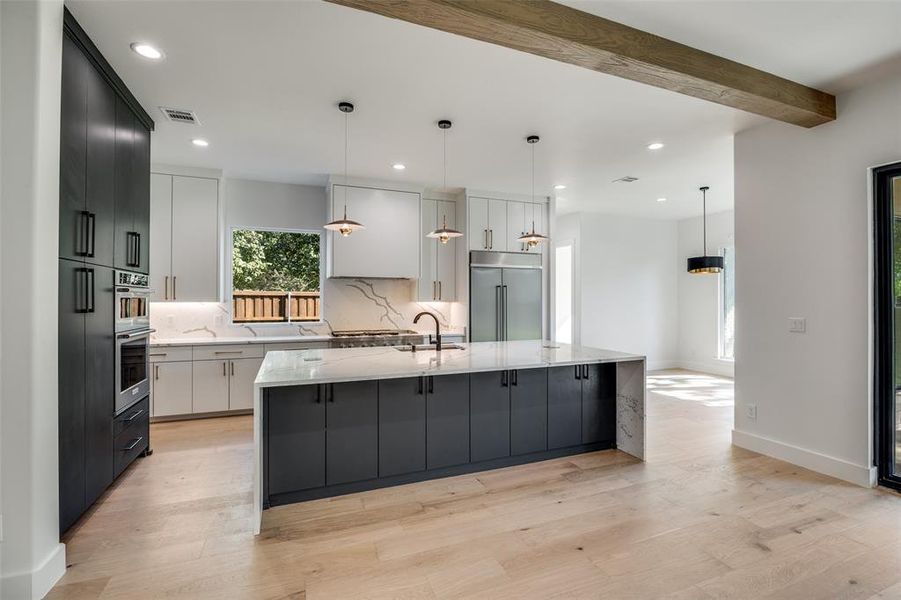 Kitchen with decorative light fixtures, a kitchen island with sink, light stone countertops, light wood-type flooring, and stainless steel appliances Kitchen with decorative light fixtures, a kitchen island with sink, light stone countertops, light wood-type flooring, and stainless steel appliances