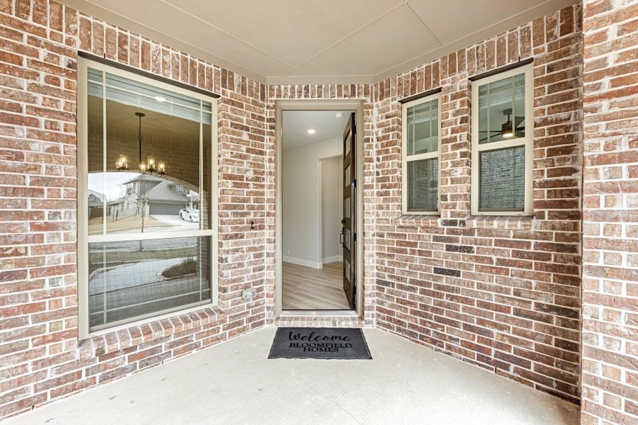 Covered front entry with brick exterior, open door, and Bloomfield Homes welcome mat Covered front entry with brick exterior, open door, and Bloomfield Homes welcome mat