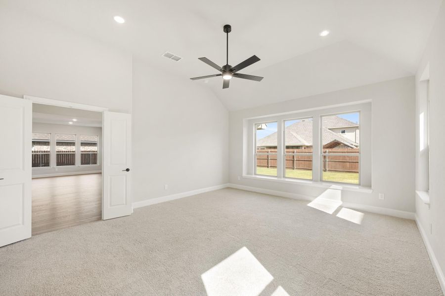 Empty bedroom with vaulted ceiling, ceiling fan, carpet flooring, and large windows overlooking a fenced backyard