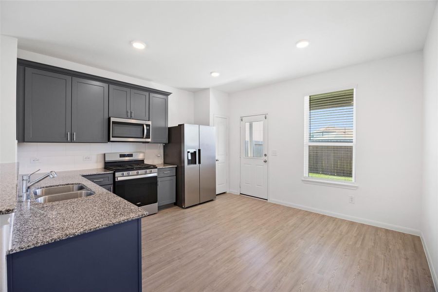kitchen features sleek dark cabinets, granite countertops, and stainless steel appliances, including a double-door refrigerator. The open space is complemented by light wood flooring and ample natural light from the window and door leading outside.