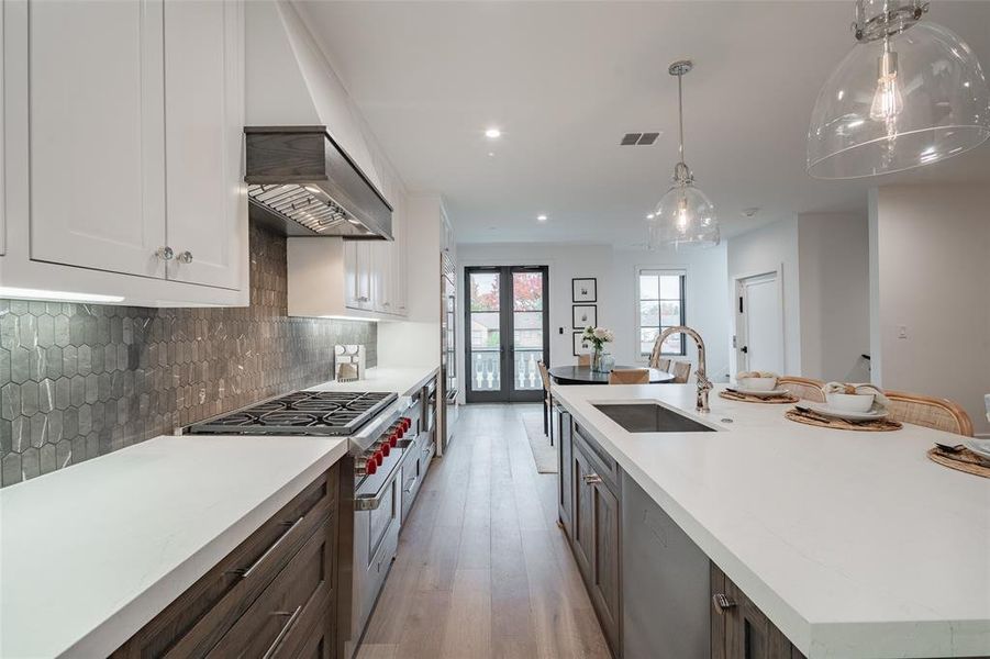 Kitchen with high end range, tasteful backsplash, white cabinets, decorative light fixtures, and custom exhaust hood