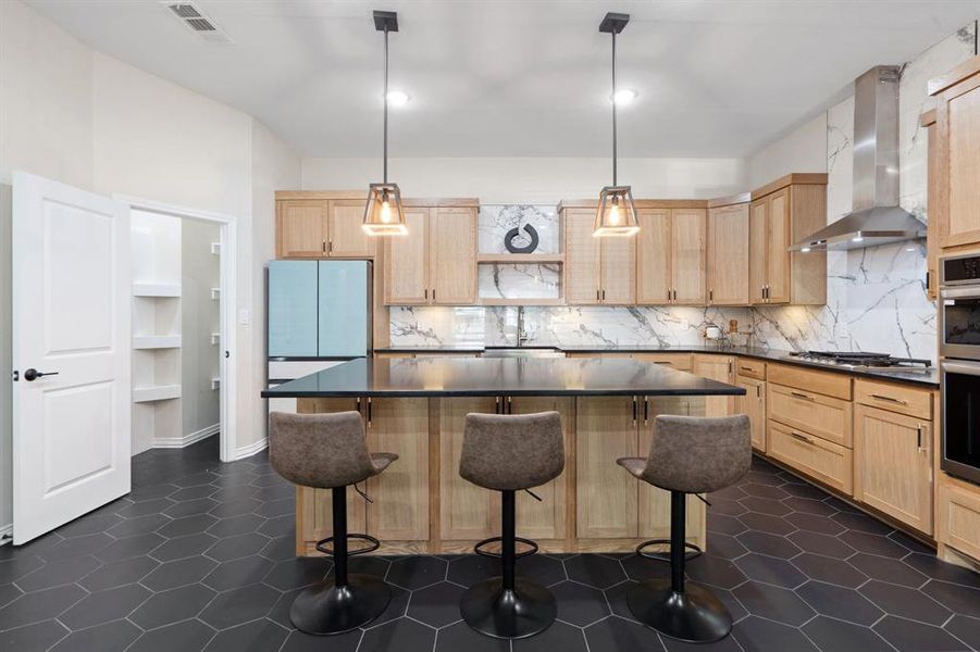 Kitchen featuring light brown cabinetry, a breakfast bar area, decorative light fixtures, a center island, and wall chimney range hood Kitchen featuring light brown cabinetry, a breakfast bar area, decorative light fixtures, a center island, and wall chimney range hood