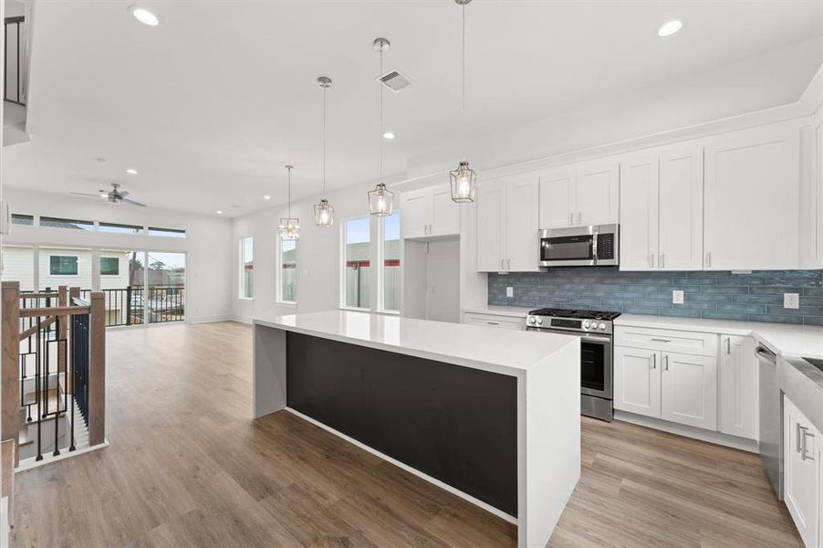 This photo showcases a modern, open-concept kitchen and living area. The kitchen features sleek white cabinetry, a large island with pendant lighting, and stainless steel appliances. The space is bright and airy, with ample natural light from large windows and contemporary wood flooring throughout.