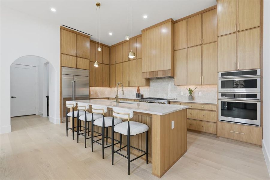 Contemporary kitchen featuring light wood-finish cabinetry, a large center island with seating, integrated stainless steel appliances, and sleek pendant lighting Contemporary kitchen featuring light wood-finish cabinetry, a large center island with seating, integrated stainless steel appliances, and sleek pendant lighting