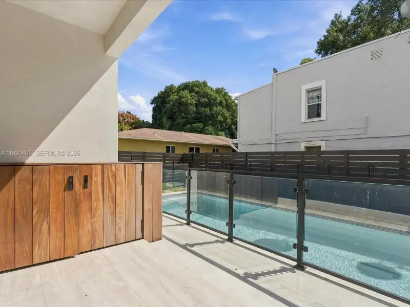 Outdoor patio featuring stone tile flooring, built-in wood-finish cabinetry, and a swimming pool enclosed by a glass-panel railing with metal frame Outdoor patio featuring stone tile flooring, built-in wood-finish cabinetry, and a swimming pool enclosed by a glass-panel railing with metal frame