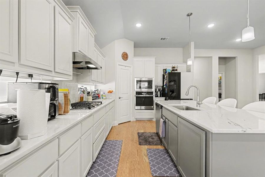 Kitchen featuring a large island with sink, white shaker-style cabinetry, light-toned countertops, built-in stainless steel appliances, and light wood-finish flooring