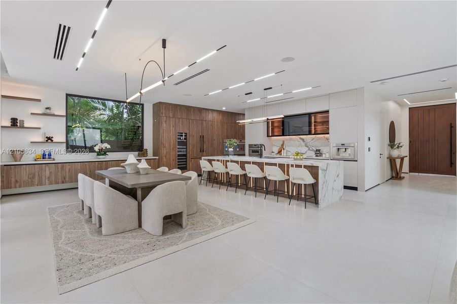 Open-concept kitchen featuring a waterfall-edge stone island, built-in panel-ready appliances, wood-finish cabinetry, linear recessed ceiling lighting, and porcelain tile flooring