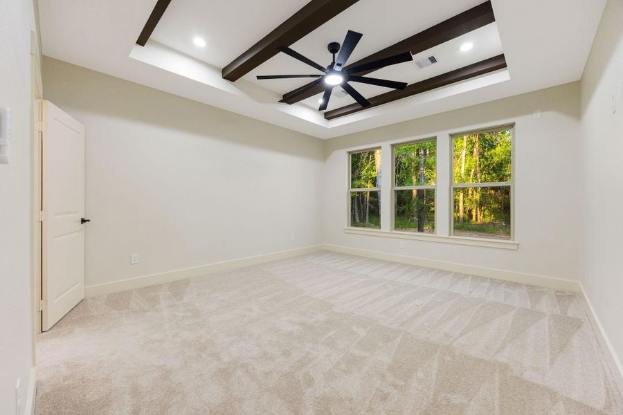 This spacious primary bedroom room features a modern design with a tray ceiling and exposed beams, a stylish ceiling fan, and large windows offering scenic views of greenery. The neutral carpet and wall colors create a warm, inviting atmosphere.