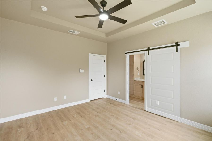 Unfurnished bedroom featuring a barn door, a tray ceiling, light wood finished floors, ensuite bath, and a ceiling fan