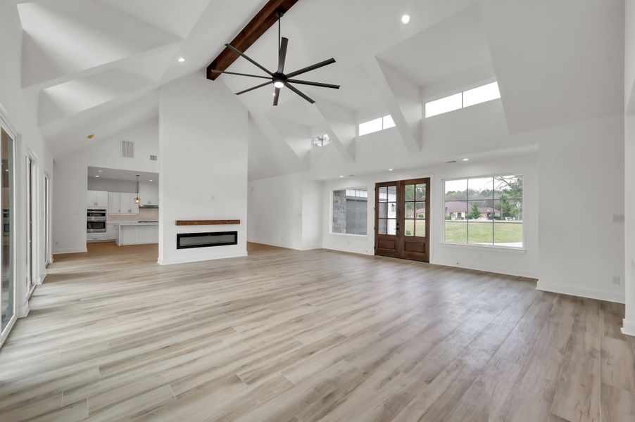 Spacious open-concept living area featuring soaring cathedral ceilings, wood-look flooring, and a striking modern ceiling fan. The double-sided electric fireplace creates a beautiful focal point while maintaining an open flow.