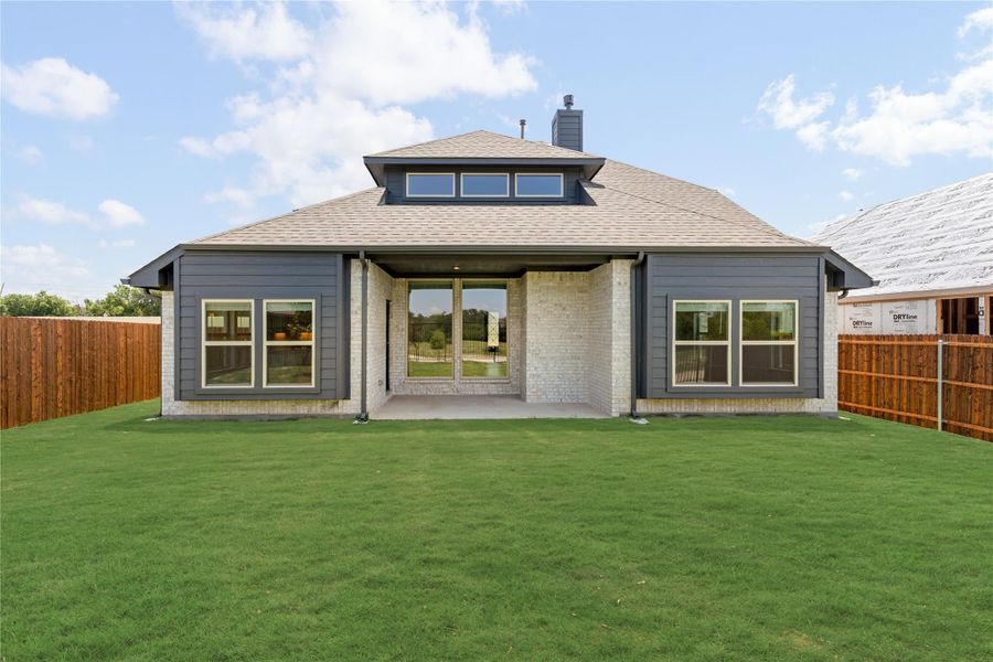 Back of single-story home with gray siding, brick accents, covered patio, and fenced green lawn. Back of single-story home with gray siding, brick accents, covered patio, and fenced green lawn.