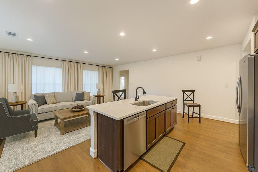 This modern open-concept living space features a cozy seating area with a neutral color palette, ample natural light, and a sleek kitchen island with a sink and stainless steel appliances.
