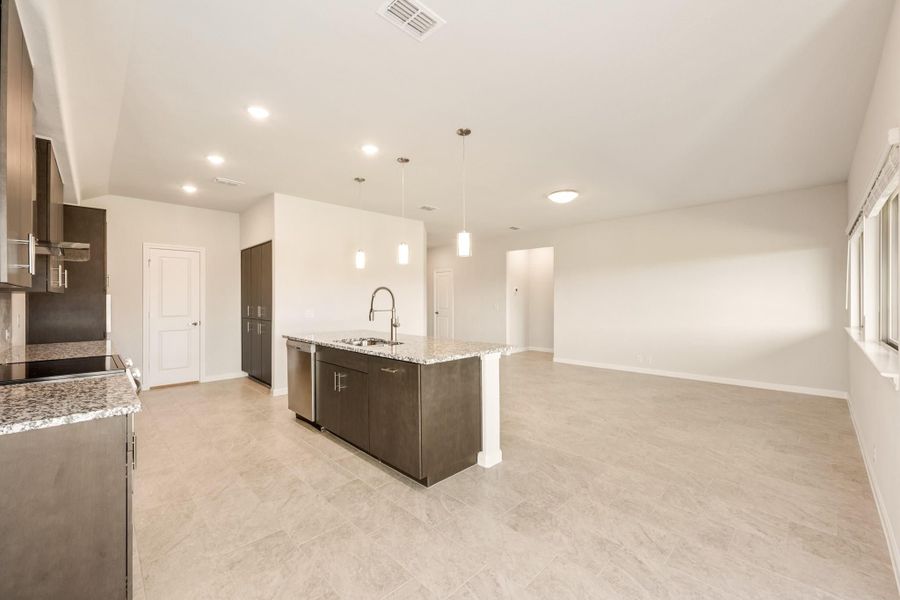 Open kitchen with dark cabinets, granite island with sink, and pendant lights opening to living area