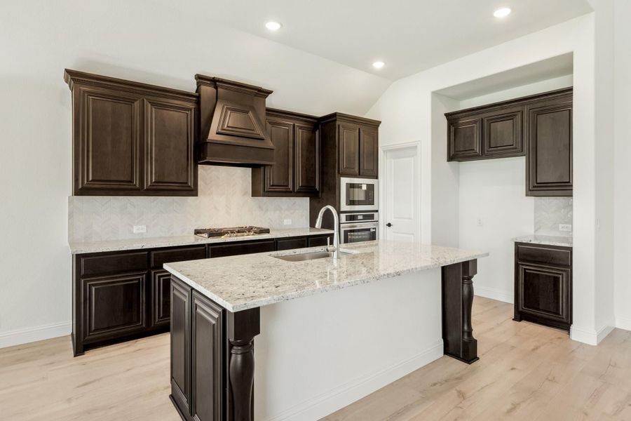 Kitchen with dark wood cabinets, granite island with sink, herringbone tile backsplash, and light hardwood floors