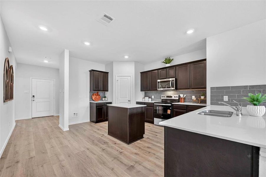 Kitchen featuring dark wood cabinetry, stainless steel appliances, a center island with a light-toned countertop, and light wood-finish flooring Kitchen featuring dark wood cabinetry, stainless steel appliances, a center island with a light-toned countertop, and light wood-finish flooring