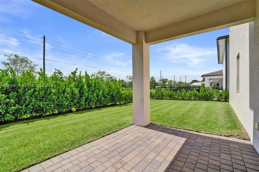 Covered patio featuring paver flooring, leading to a manicured lawn and mature privacy hedges